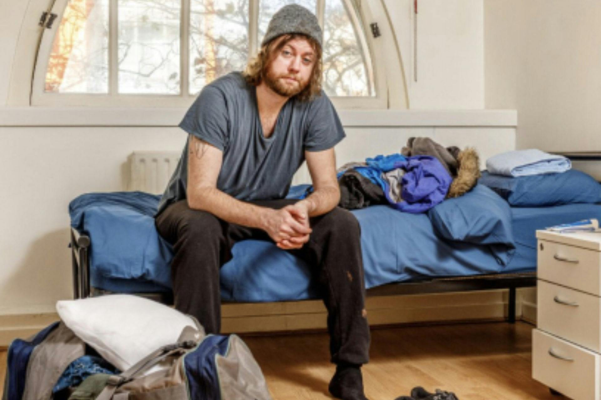 A man with a beard and beanie hat is sitting on the edge of a bed